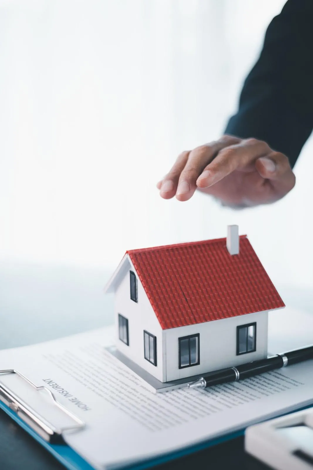 A hand held above a small model house resting on a home insurance document